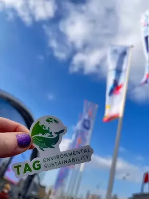 Photo of TAG Environmental Sustainability logo sticker with KubeCon+CloudNativeCon EU flags in the background